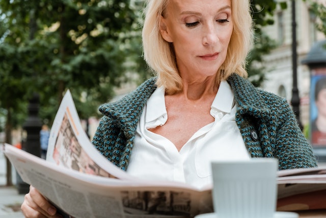 A landlord with a blond bob haircut sits at an outdoor cafe and reviews for rent ads in the classified section of a newspaper